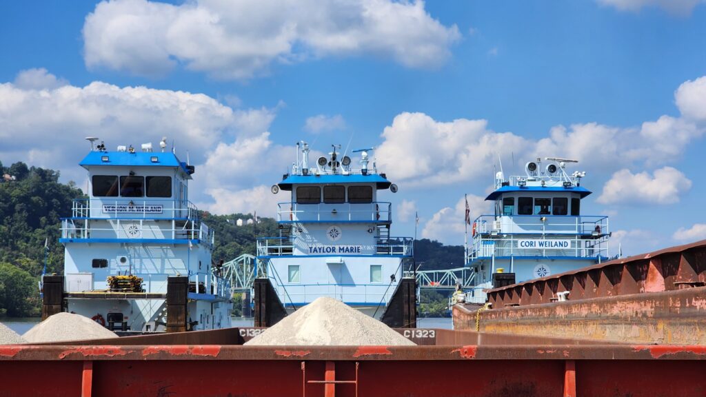Three Borghese Lane towboats moored side by side on a bright day with piled clouds, seen across the coamings of a red hopper barge holding two cones of sand.