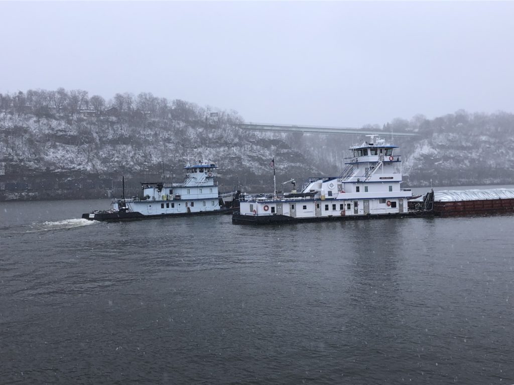 Two towboats passing in heavy snowfall on a grey river, one light and one pushing a line of covered hopper barges, with a snow-covered hillside behind.