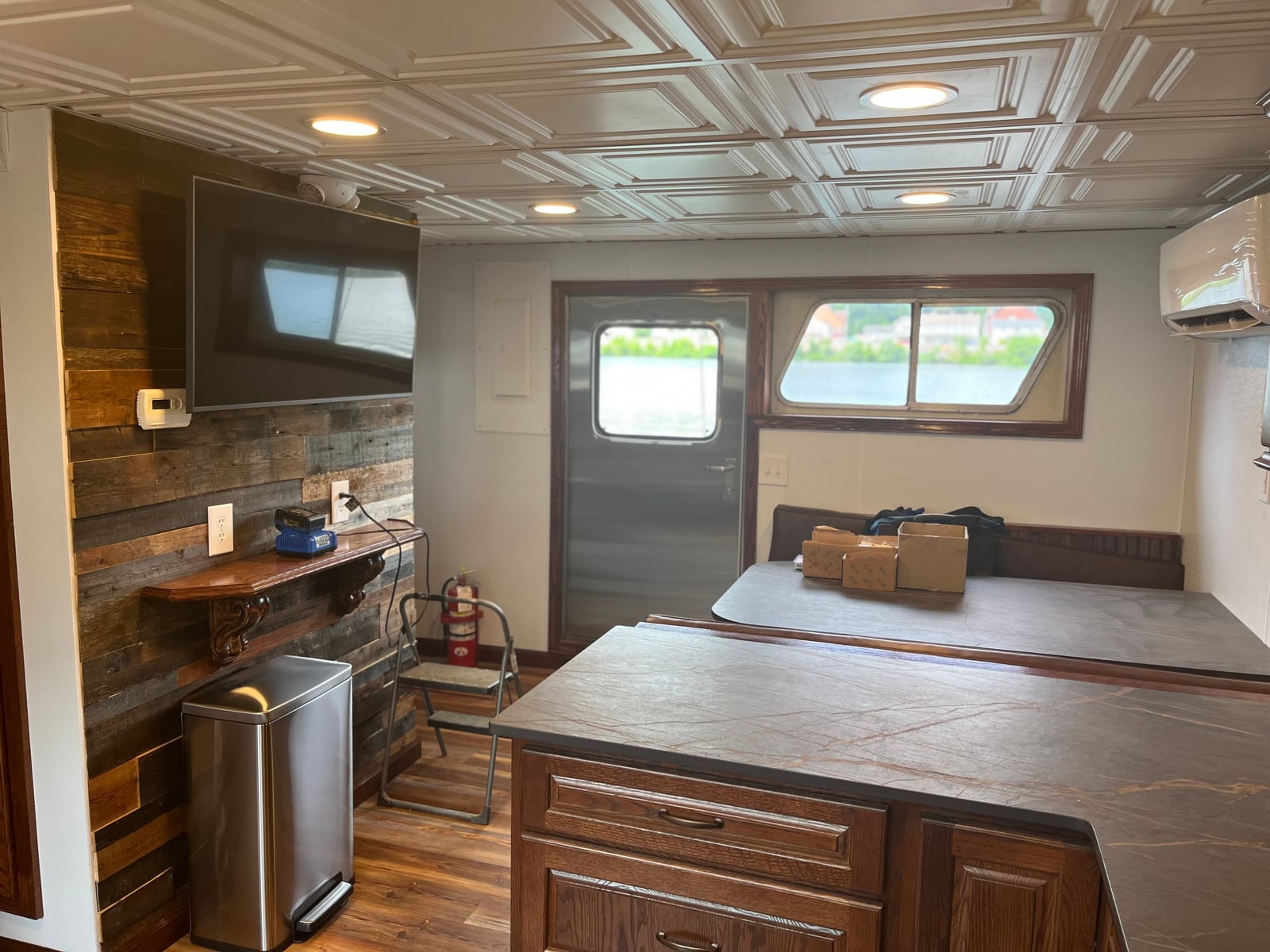 A towboat galley seating area beside wood cabinets, a wall-mounted television and a metal waste bin.
