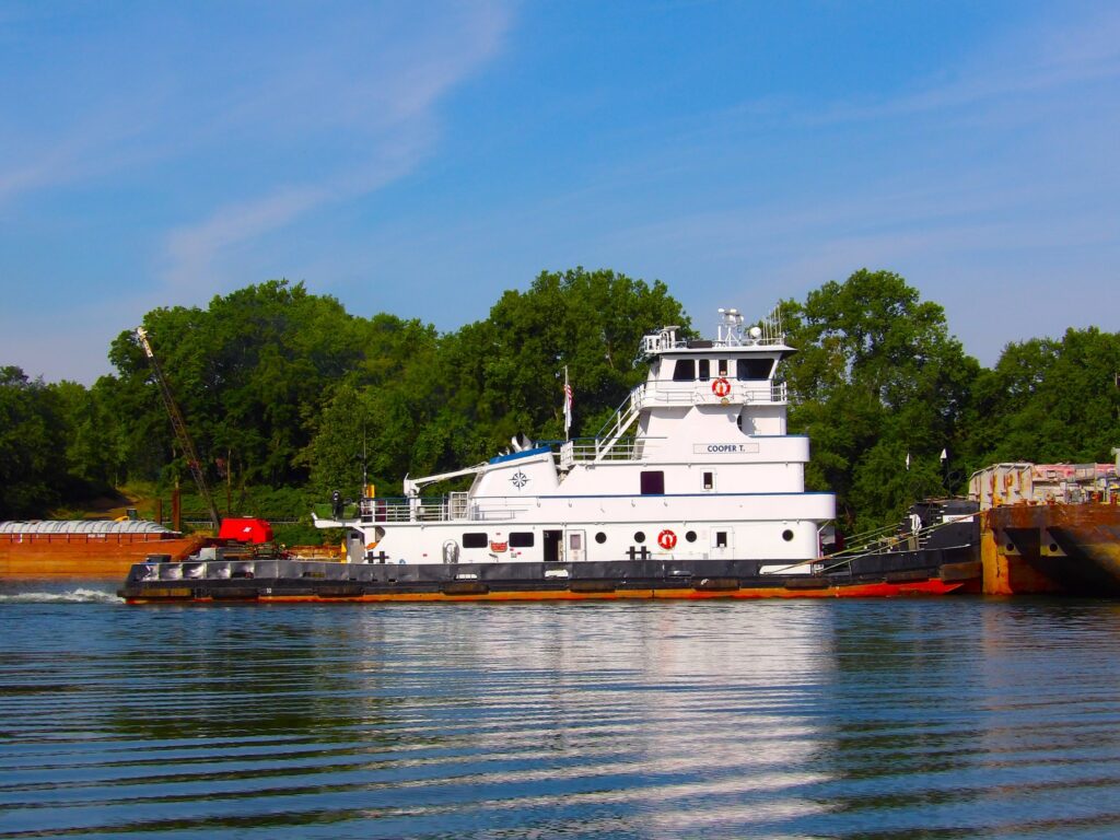 The towboat Cooper T. alongside a loaded hopper barge on a clear summer day, green trees on the far bank.