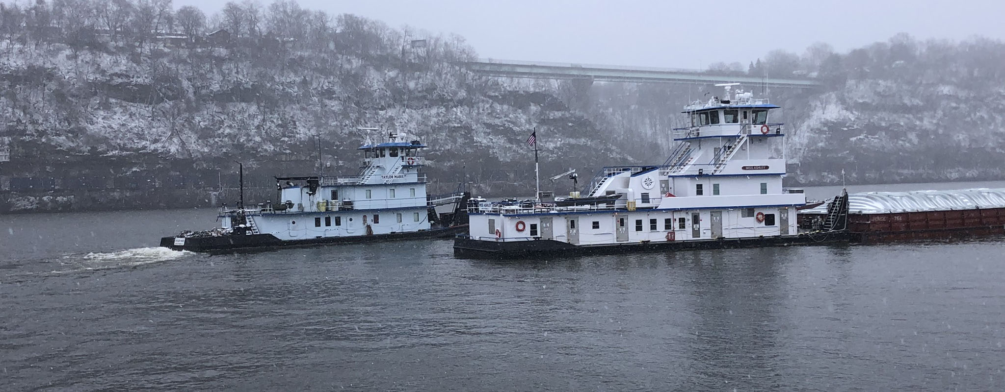 Two towboats working in falling snow on a grey river below a snow-dusted cliff, one pushing a covered hopper barge, a highway bridge in the haze behind them.