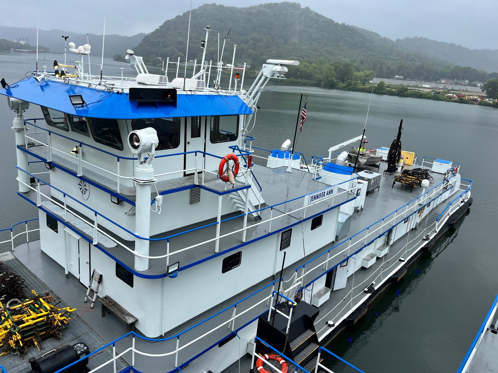 Jennifer Ann viewed from above, showing its blue pilothouse roof, white decks and stern against a wooded riverbank.