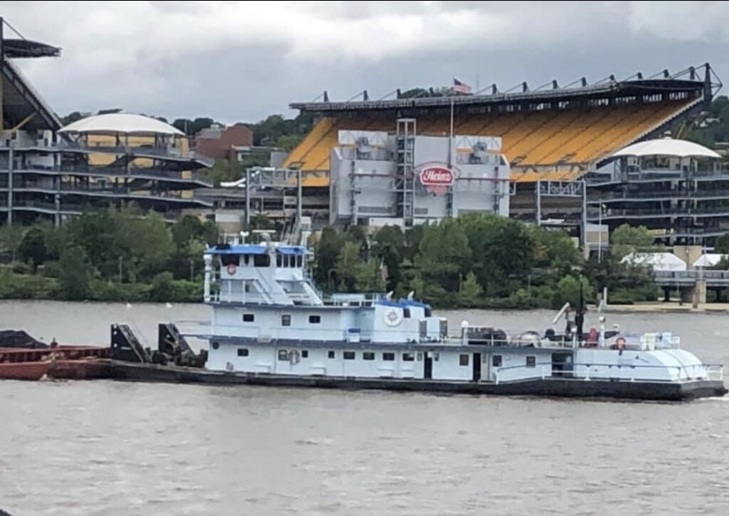A towboat pushing an empty red hopper barge past Acrisure Stadium on the Ohio River at Pittsburgh under a flat grey sky.