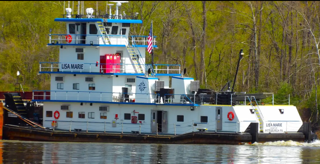 The towboat Lisa Marie, a three-deck boat in white with blue trim, moored against a bare-tree bank in early spring. LISA MARIE is lettered on the pilothouse deck and again on the stern above PORT OF PITTSBURGH PA.