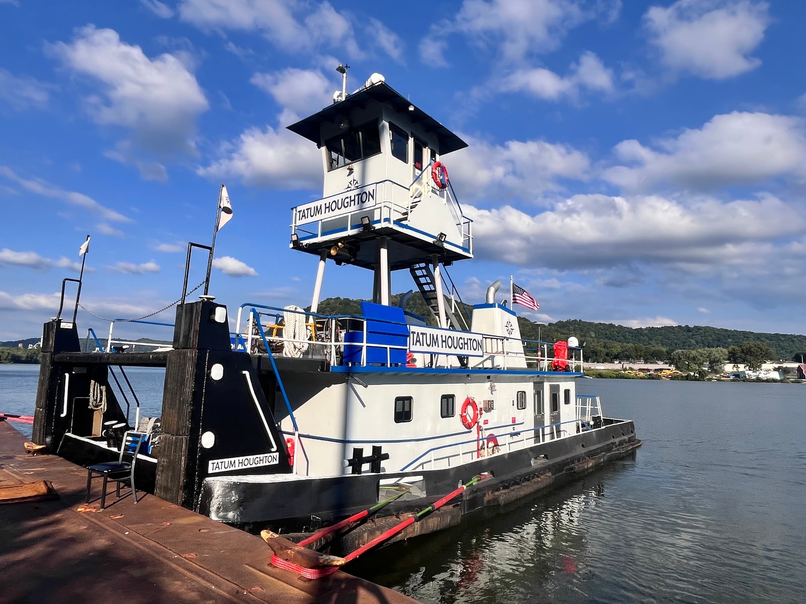 Tatum Houghton moored beside a barge under a blue sky, with its name visible on the tall pilothouse.